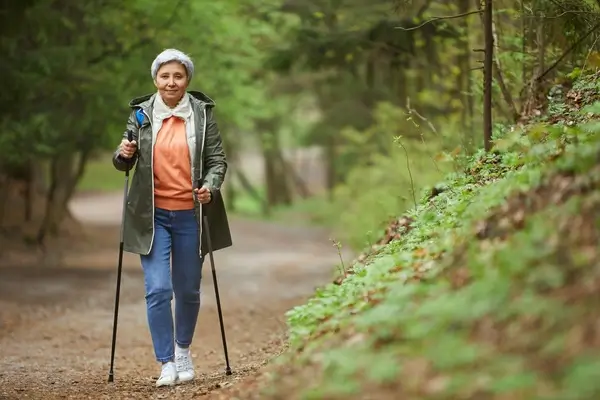 An older woman walks through a forest with hiking sticks