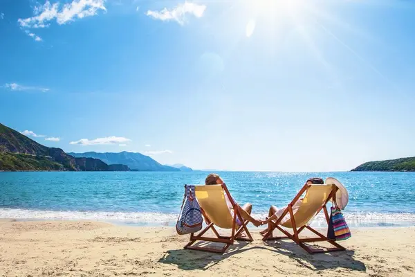 Two people relax on deckchairs on holiday on a beach under a bright blue, sunny sky